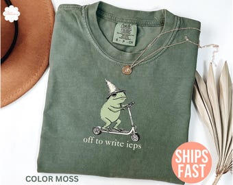 May include: Green Comfort Colors t-shirt with a whimsical frog illustration wearing a wizard hat and riding a scooter. The text "off to write ieps" is printed below the frog. The shirt is color moss.