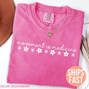 May include: A pink t-shirt with the phrase "movement is medicine" in white script and daisy graphics. A gold necklace rests on the shirt. The shirt is labeled "Comfort Colors" and the color is "Crushberry".