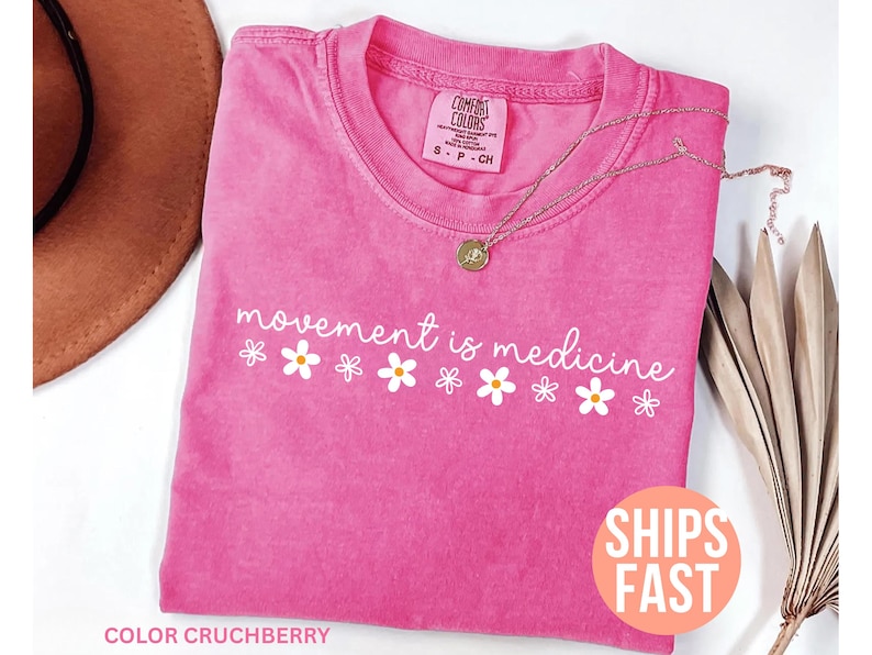 May include: A pink t-shirt with the phrase "movement is medicine" in white script and daisy graphics. A gold necklace rests on the shirt. The shirt is labeled "Comfort Colors" and the color is "Crushberry".