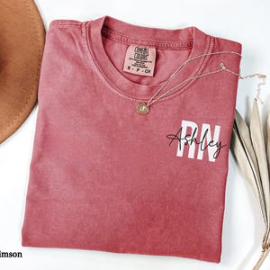 May include: Crimson red t-shirt with a white print that says "Ashley RN".