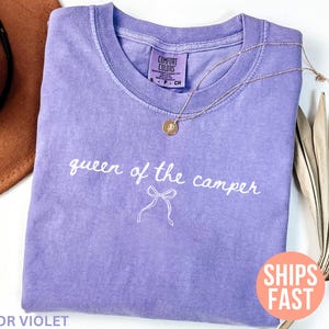 May include: A lavender Comfort Colors t-shirt with the phrase "queen of the camper" in white script. A gold necklace with a pendant rests on the shirt. The words "Color Violet" are at the bottom.