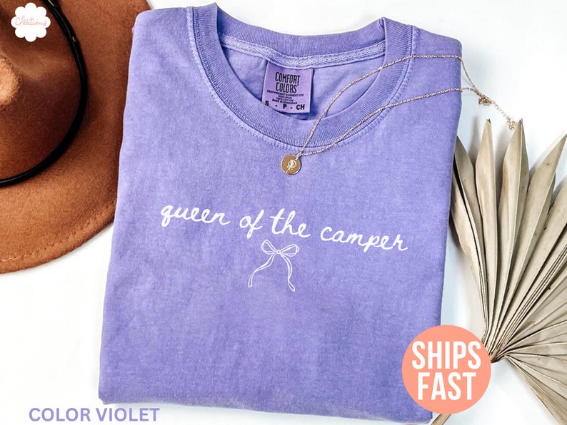May include: A lavender Comfort Colors t-shirt with the phrase "queen of the camper" in white script. A gold necklace with a pendant rests on the shirt. The words "Color Violet" are at the bottom.