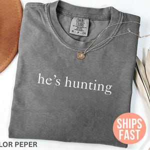 May include: A gray Comfort Colors t-shirt with the phrase "he's hunting" printed in white. A gold pendant necklace rests on the shirt. A brown hat is partially visible in the upper left corner.
