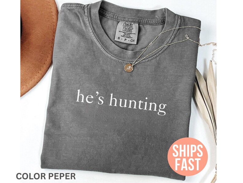 May include: A gray Comfort Colors t-shirt with the phrase "he's hunting" printed in white. A gold pendant necklace rests on the shirt. A brown hat is partially visible in the upper left corner.