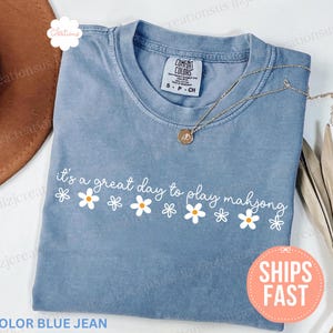 May include: A blue jean colored t-shirt with the phrase "it's a great day to play mahjong" in white script, accented with white daisy graphics. A gold necklace and a brown hat are also visible.