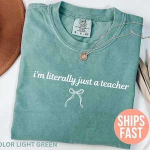 May include: Light green t-shirt with the text "i'm literally just a teacher" in white. A silver necklace with a pendant is on the shirt. The shirt is folded and has a tag that says "Comfort Colors".