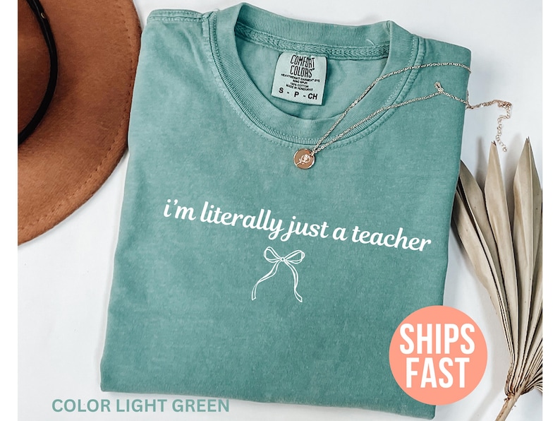 May include: Light green t-shirt with the text "i'm literally just a teacher" in white. A silver necklace with a pendant is on the shirt. The shirt is folded and has a tag that says "Comfort Colors".