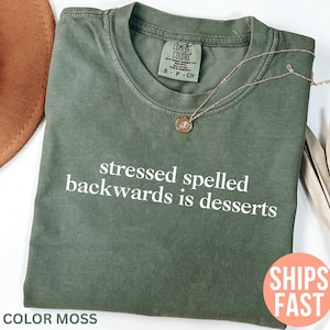 May include: A moss green t-shirt with the slogan "stressed spelled backwards is desserts" in white. The shirt has a crew neck and short sleeves. A gold necklace with a pendant rests on the shirt.