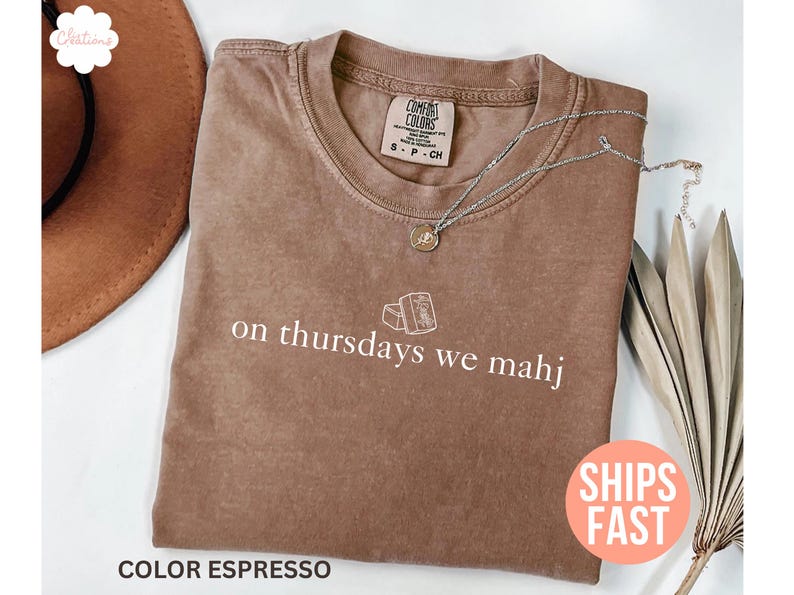 以下が含まれることがあります： エスプレッソカラーのComfort Colors Tシャツ。白い文字で「on thursdays we mahj」と書かれています。ペンダント付きのシルバーネックレスがシャツの上にあります。シャツは白い面に折りたたまれています。