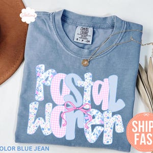Könnte beinhalten: Blaues Jeans-T-Shirt mit den Worten "Postal Worker" in einem floralen und gestreiften Design. Eine rosa Schleife befindet sich in der Mitte des Textes. Das Shirt hat einen Rundhalsausschnitt und kurze Ärmel. Der Text "Ships Fast" steht in einem Pfirsichkreis.
