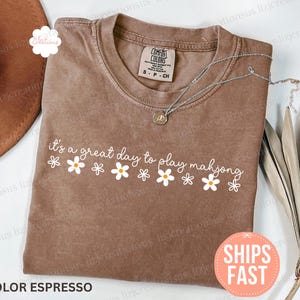 May include: Espresso-colored t-shirt with the text "It's a great day to play mahjong" in white, decorated with daisy graphics. A silver necklace with a pendant is on the shirt. The shirt is folded on a surface with a brown hat.
