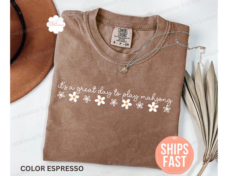 May include: Espresso-colored t-shirt with the text "It's a great day to play mahjong" in white, decorated with daisy graphics. A silver necklace with a pendant is on the shirt. The shirt is folded on a surface with a brown hat.