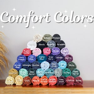 May include: A display of rolled-up t-shirts in various colors, including white, black, blue, and green. The image features the text "Comfort Colors" in a decorative font, with each shirt labeled with its color name.