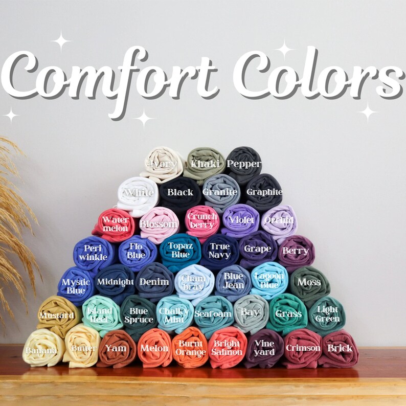 May include: A display of rolled-up t-shirts in various colors, including white, black, blue, and green. The image features the text "Comfort Colors" in a decorative font, with each shirt labeled with its color name.