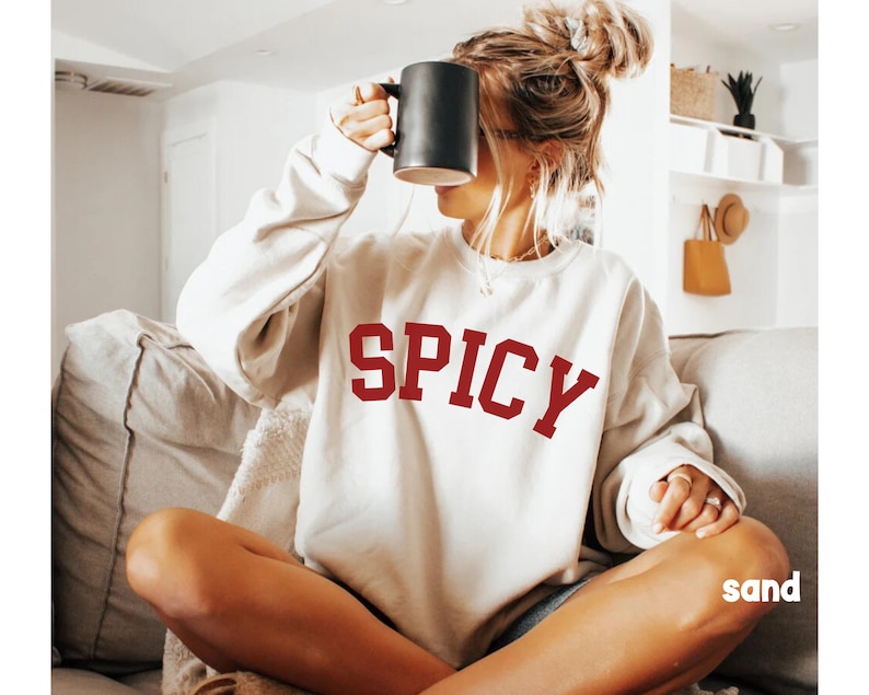Peut inclure: Un sweat-shirt blanc avec le mot "SPICY" imprim&eacute; en rouge sur le devant. Le sweat-shirt est port&eacute; par une personne assise sur un canap&eacute;.
