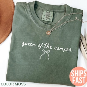 May include: A moss green Comfort Colors t-shirt with the phrase "queen of the camper" in white script. A gold necklace with a pendant rests on the shirt. A brown hat is partially visible.