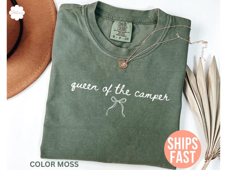 May include: A moss green Comfort Colors t-shirt with the phrase "queen of the camper" in white script. A gold necklace with a pendant rests on the shirt. A brown hat is partially visible.