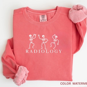 May include: Coral-colored sweatshirt with white graphic of three dancing skeletons and the word "RADIOLOGY" below. The sleeves are rolled up, revealing a lighter pink lining. The sweatshirt is a Comfort Colors brand.