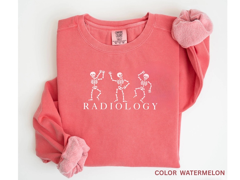 May include: Coral-colored sweatshirt with white graphic of three dancing skeletons and the word "RADIOLOGY" below. The sleeves are rolled up, revealing a lighter pink lining. The sweatshirt is a Comfort Colors brand.
