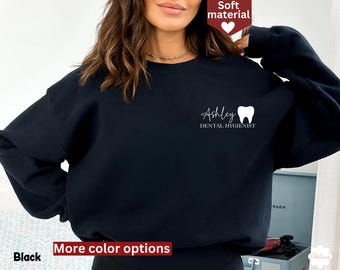 May include: Black sweatshirt with the name "Ashley" and a tooth graphic, along with the words "Dental Hygienist" in white script. The sweatshirt is made of a soft material. The words "More color options" and the word "Black" are also visible.