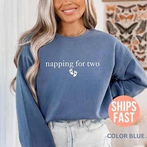 May include: Blue jean-colored sweatshirt with the text "napping for two" and two small footprints. The sweatshirt is a long-sleeved crew neck. The image also includes a round sticker that says "SHIPS FAST".