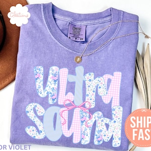 May include: A violet Comfort Colors t-shirt with the words "ULTRA SOUND" in a playful font. The letters are decorated with floral and gingham patterns, and a pink bow. A gold necklace rests on the shirt.