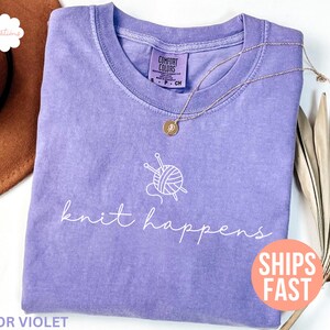 May include: A violet Comfort Colors t-shirt with the phrase "knit happens" in white script. A graphic of yarn and knitting needles is above the text. A gold necklace with a pendant is on the shirt. The shirt is folded.