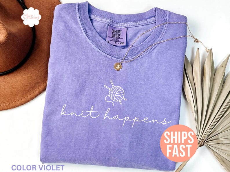 May include: A violet Comfort Colors t-shirt with the phrase "knit happens" in white script. A graphic of yarn and knitting needles is above the text. A gold necklace with a pendant is on the shirt. The shirt is folded.