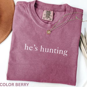 May include: Berry-colored t-shirt with the text "he's hunting" in white. A gold-colored necklace with a pendant is draped over the shirt. The shirt is a Comfort Colors brand.