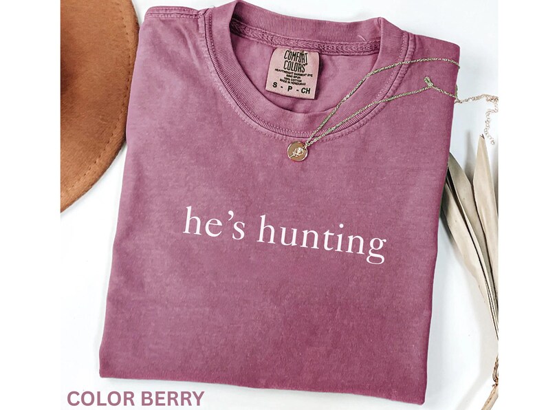 May include: Berry-colored t-shirt with the text "he's hunting" in white. A gold-colored necklace with a pendant is draped over the shirt. The shirt is a Comfort Colors brand.