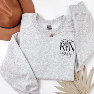 May include: A gray sweatshirt with a black print of a medical caduceus symbol and the letters "RN" with the name "Ashley" underneath.