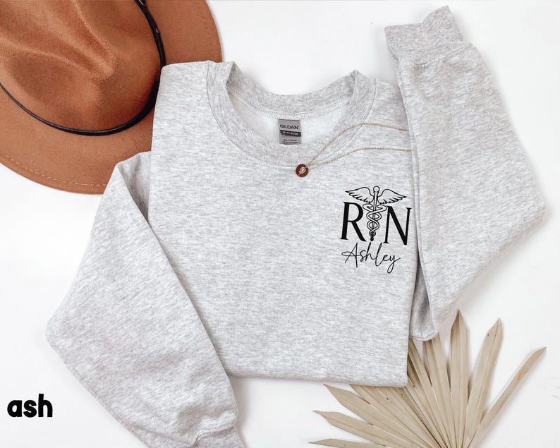 May include: A gray sweatshirt with a black print of a medical caduceus symbol and the letters "RN" with the name "Ashley" underneath.