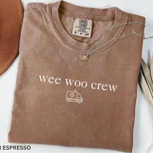 May include: Espresso-colored t-shirt with the text "wee woo crew" and an ambulance graphic. The shirt has a crew neck and short sleeves. A silver necklace with a pendant is draped across the shirt.