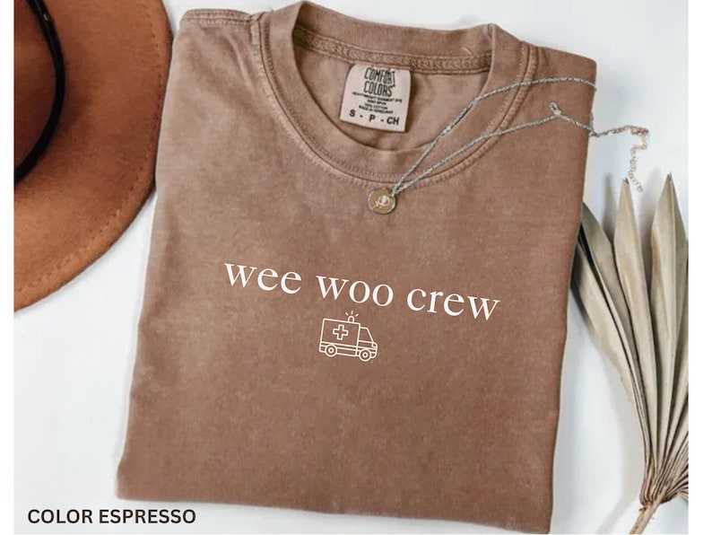 May include: Espresso-colored t-shirt with the text "wee woo crew" and an ambulance graphic. The shirt has a crew neck and short sleeves. A silver necklace with a pendant is draped across the shirt.