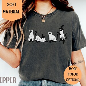 May include: Pepper-colored t-shirt featuring a playful Halloween design. The graphic depicts four cartoon cats dressed as ghosts, with one wearing a witch hat. The shirt has the text "SOFT MATERIAL" and "MORE COLOR OPTIONS".