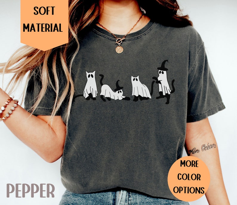 May include: Pepper-colored t-shirt featuring a playful Halloween design. The graphic depicts four cartoon cats dressed as ghosts, with one wearing a witch hat. The shirt has the text "SOFT MATERIAL" and "MORE COLOR OPTIONS".