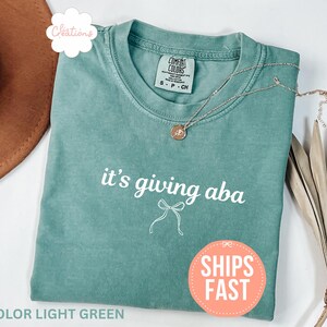 May include: A light green crew neck t-shirt featuring the white text it's giving aba and a small white line drawing of a bow centered below the text. The shirt has a Comfort Colors brand label at the inner neckline and a short silver necklace.