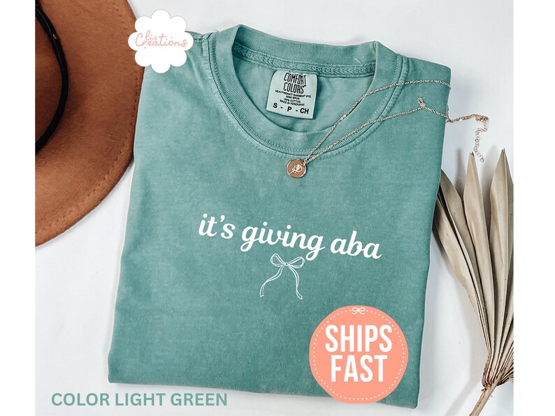 May include: A light green crew neck t-shirt featuring the white text it's giving aba and a small white line drawing of a bow centered below the text. The shirt has a Comfort Colors brand label at the inner neckline and a short silver necklace.