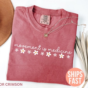 May include: Crimson-colored t-shirt with the phrase "movement is medicine" in white script and daisy graphics. A silver necklace with a pendant rests on the shirt. The shirt is folded and displayed with a hat and dried plant.