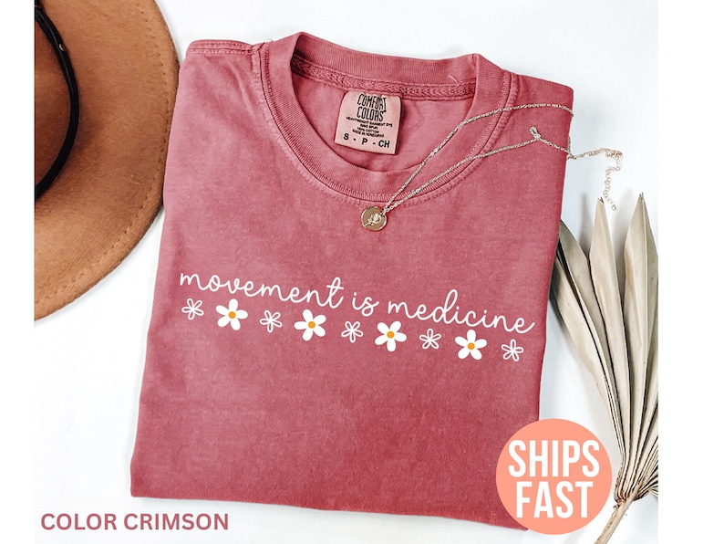 May include: Crimson-colored t-shirt with the phrase "movement is medicine" in white script and daisy graphics. A silver necklace with a pendant rests on the shirt. The shirt is folded and displayed with a hat and dried plant.