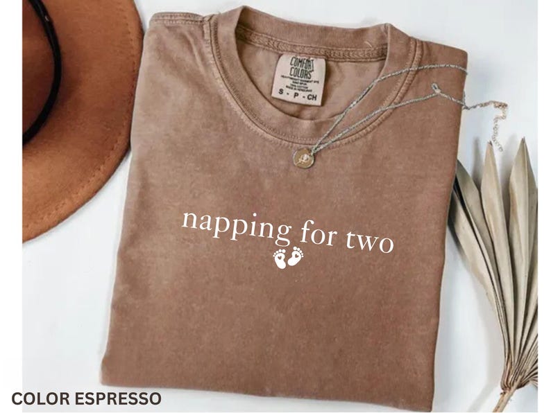 Puede incluir: Camiseta color espresso con el texto "napping for two" y dos peque&ntilde;as huellas. Un collar plateado con un colgante descansa sobre la camiseta. Un sombrero marr&oacute;n es visible en el fondo.
