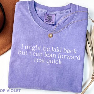 May include: A lavender Comfort Colors t-shirt with the text "i might be laid back but i can lean forward real quick" in white. A gold necklace and a brown hat are also visible.