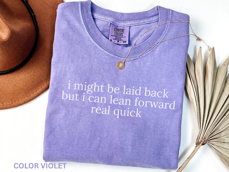 May include: A lavender Comfort Colors t-shirt with the text "i might be laid back but i can lean forward real quick" in white. A gold necklace and a brown hat are also visible.