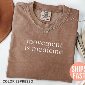 K&ouml;nnte beinhalten: Espressofarbenes T-Shirt mit dem wei&szlig;en Schriftzug &bdquo;movement is medicine&ldquo;. Das Shirt hat einen Rundhalsausschnitt und ein kleines Etikett. Eine silberne Halskette mit Anh&auml;nger liegt auf dem Shirt. Ein brauner Hut und getrocknete Pflanzen sind im Hintergrund.