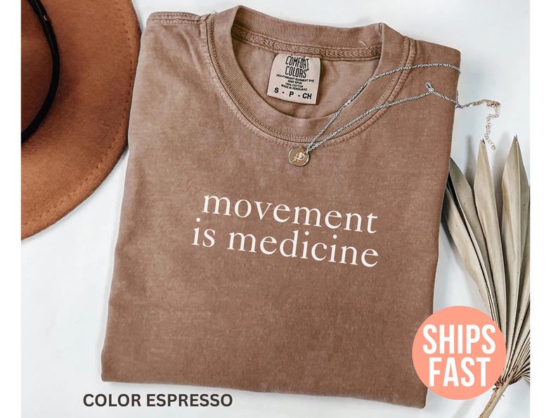 K&ouml;nnte beinhalten: Espressofarbenes T-Shirt mit dem wei&szlig;en Schriftzug &bdquo;movement is medicine&ldquo;. Das Shirt hat einen Rundhalsausschnitt und ein kleines Etikett. Eine silberne Halskette mit Anh&auml;nger liegt auf dem Shirt. Ein brauner Hut und getrocknete Pflanzen sind im Hintergrund.