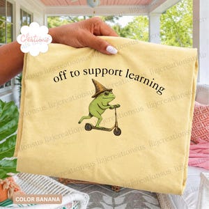 Puede incluir: Una camiseta color amarillo banana con un gráfico de una rana verde que lleva un sombrero puntiagudo marrón y monta un patinete. Sobre la rana, el texto off to support learning aparece en una fuente serif negra arqueada sobre la tela.