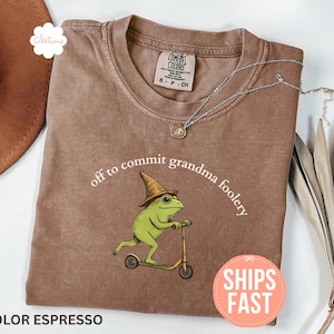 以下が含まれることがあります： エスプレッソカラーのTシャツ。スクーターに乗ったカエルの風変わりなグラフィックと魔女の帽子が描かれています。「off to commit grandma foolery」の文字がアーチ状に配置されています。シルバーのネックレスと帽子が見えます。