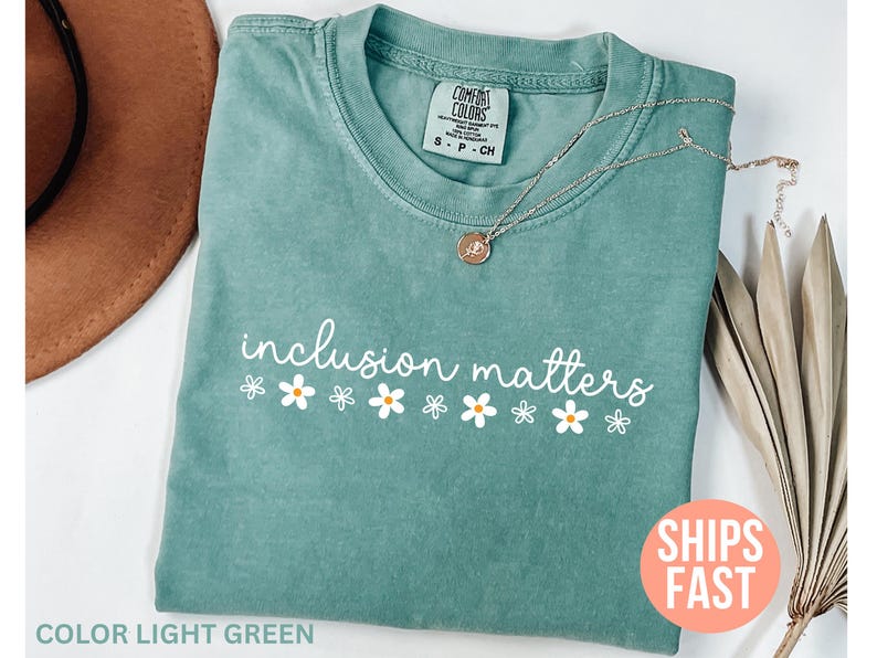 May include: A light green t-shirt with the words "inclusion matters" in white script and daisy accents. A silver necklace with a pendant rests on the shirt. The shirt is folded, and a brown hat is visible in the background.