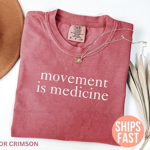 K&ouml;nnte beinhalten: Karmesinrotes T-Shirt mit dem wei&szlig;en Text "movement is medicine". Eine goldene Halskette mit Anh&auml;nger liegt auf dem Shirt. Das Shirt ist Teil einer Bekleidungskollektion.