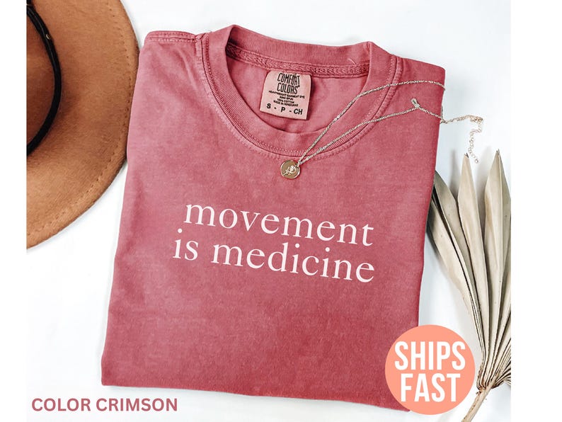 K&ouml;nnte beinhalten: Karmesinrotes T-Shirt mit dem wei&szlig;en Text "movement is medicine". Eine goldene Halskette mit Anh&auml;nger liegt auf dem Shirt. Das Shirt ist Teil einer Bekleidungskollektion.